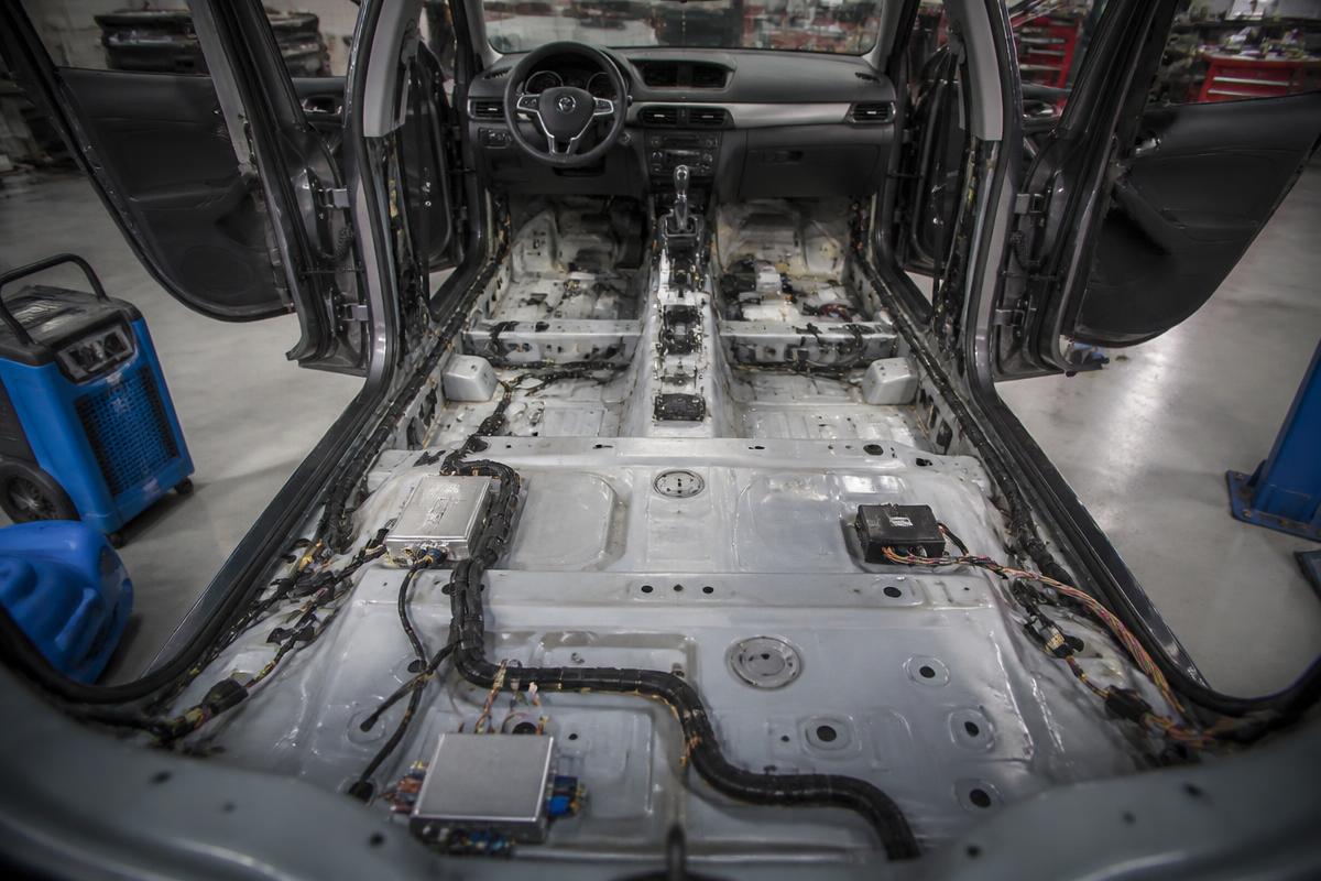 Vehicle interior stripped to the bare floor pan with carpet and seats removed, exposing wiring and floor modules for flood damage inspection